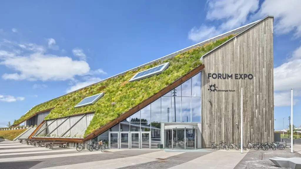 Stor byggning med trekledning og glassfasade merket «FORUM EXPO». Et skrått tak er dekket av sedum i kassetter/grønt tak, med to takvinduer. Sykler står ved inngangen, og himmelen er blå med spredte skyer.