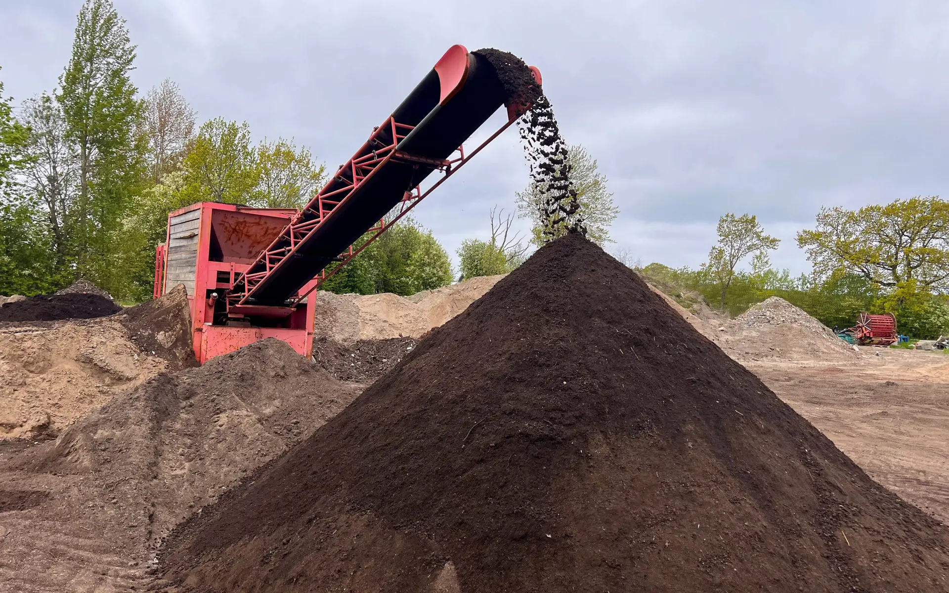 Sikteanlegg som sorterer vekstjord via transportbånd, der jord faller ned og bygger en stor haug på et gruslagt område utendørs.