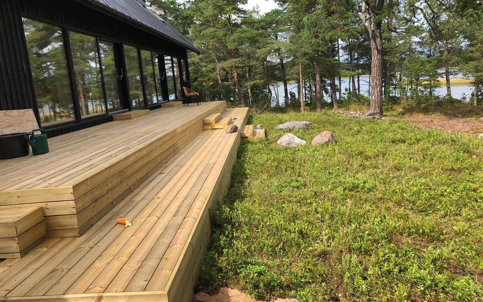 Treterrasse ved en moderne, mørk hytte med store vindusflater. Foran terrassen ligger et naturlig lyngdekke med noen steiner, furutrær og utsikt mot vann i bakgrunnen.
