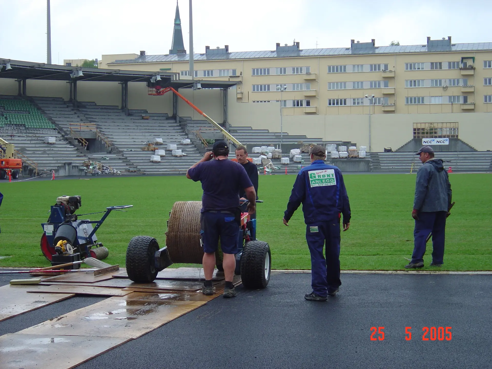 Arbeidere legger ferdigplen på Bislett stadion i 2005: en stor grønn gressflate inne på banen, mens flere personer står ved kanten med utstyr og en vogn med rull. Tribuner og bygge-/anleggsarbeid ses i bakgrunnen, og datostempel «25 5 2005» står nederst til høyre.
