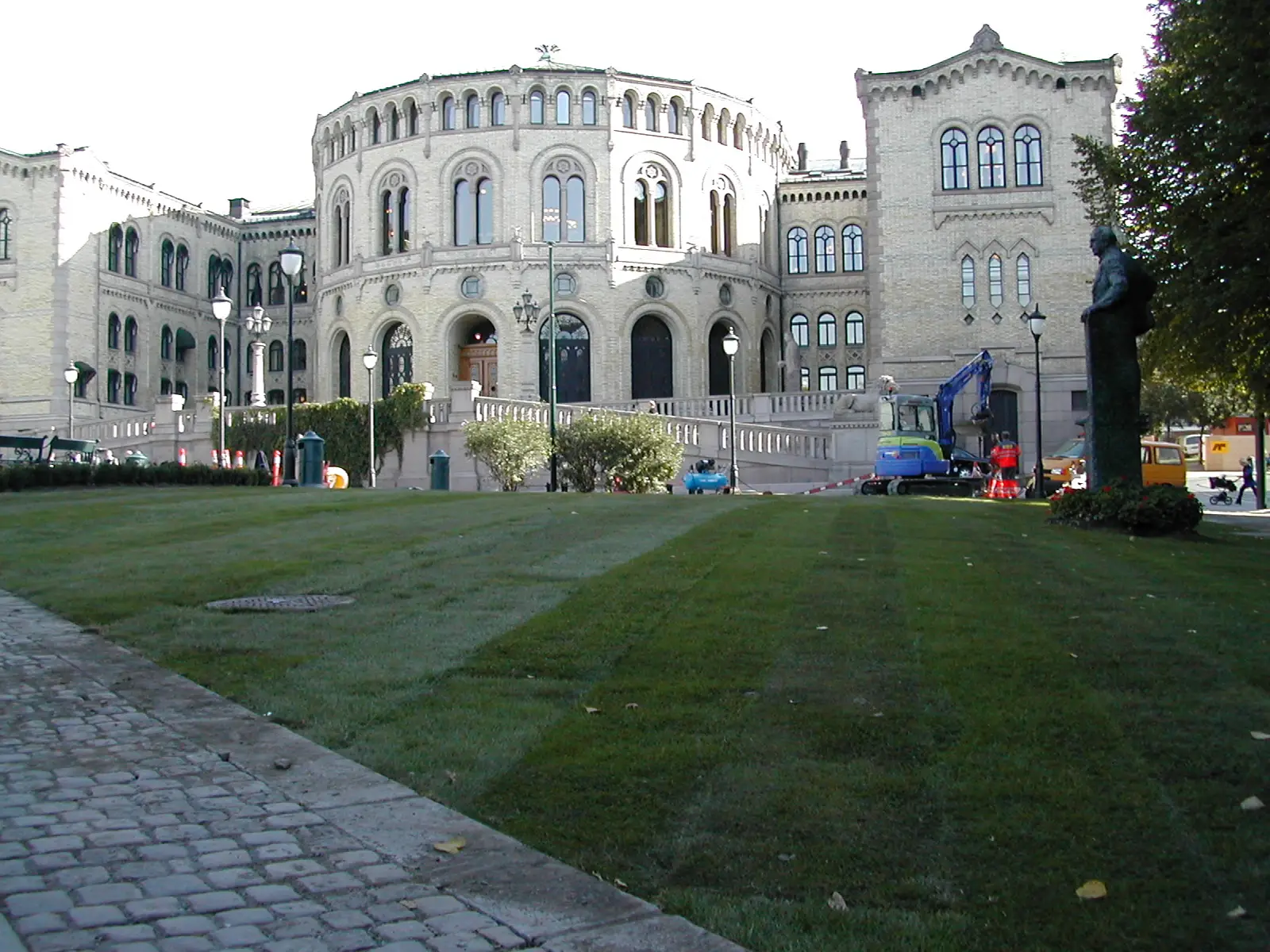Ferdigplen ligger nyanlagt foran Stortinget: brede, grønne gressflater med tydelige skjøter/striper, brosteinskant i forgrunnen og det lyse mursteinsbygget med buede vinduer i bakgrunnen. Til høyre sees anleggsmaskin og sperringer ved området.