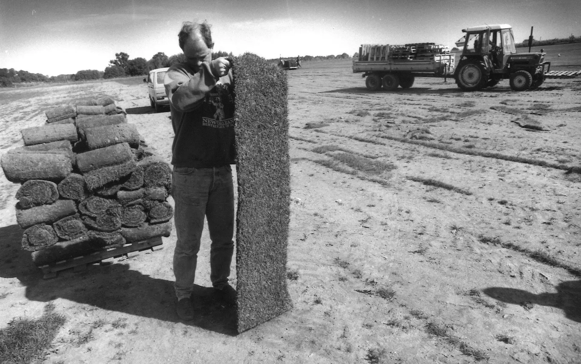 Svart-hvitt arkivfoto fra gressproduksjon: en mann står på et jorde og holder opp en lang remse ferdigplen. Ved siden av ligger en pall med rullede gressruller, og i bakgrunnen står en traktor med henger på jordet.