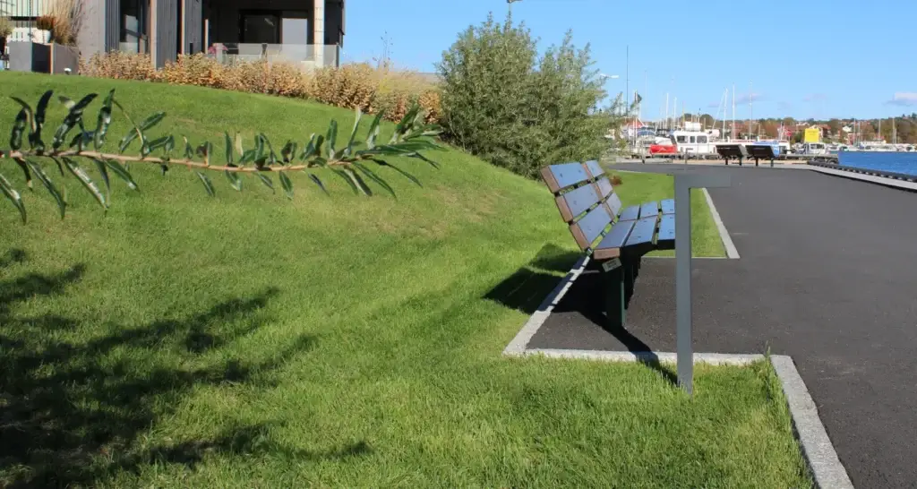 Grønn ferdigplen i en skråning ved en asfaltert gangvei med benk, med båthavn og bygninger i bakgrunnen på en solrik dag.