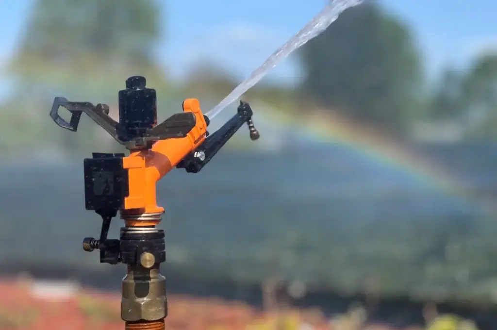 Nærbilde av en oransje og svart vannspreder i et automatisk vanningssystem som spruter en vannstråle. I bakgrunnen er det uskarpt landskap med antydning til en regnbue i vannspruten.