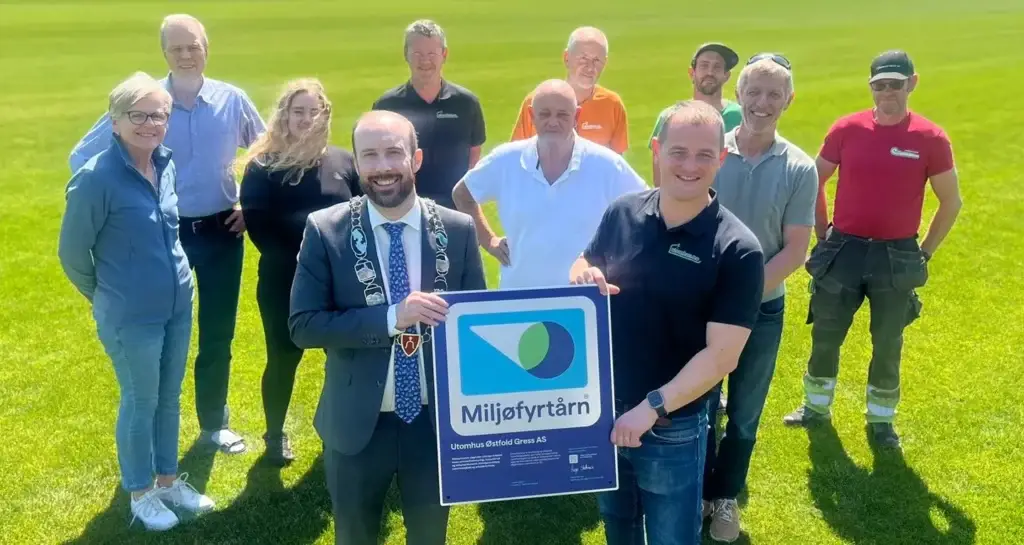 Miljøfyrtårn-sertifiserinGruppebilde utendørs på en grønn gresslette: ordføreren i dress med ordførerkjede står sammen med flere personer og holder et stort «Miljøfyrtårn»-diplom/skilt foran seg, mens resten av gruppen smiler i bakgrunnen i solskinn.g diplom overrekkelse av ordføreren i Moss