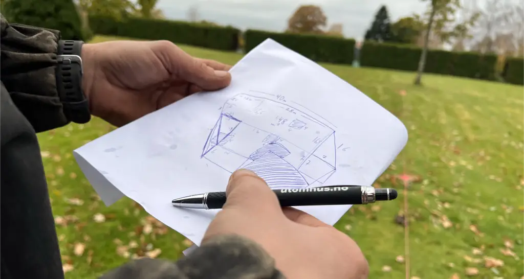 Nærbilde av to hender som holder et ark med en håndtegnet skisse og mål på hagen. En penn peker på tegningen. I bakgrunnen ses en gressplen med litt løv og oppmerkingssnor i bakken, med hekk og trær i det fjerne.