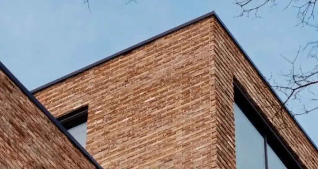 Nærbilde av hjørnet på et moderne mursteinsbygg sett nedenfra, med store vindusflater og blå himmel i bakgrunnen, samt noen nakne greiner øverst i bildet.