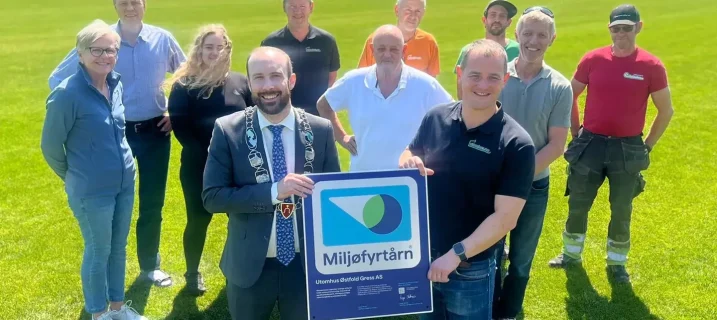 Miljøfyrtårn-sertifiserinGruppebilde utendørs på en grønn gresslette: ordføreren i dress med ordførerkjede står sammen med flere personer og holder et stort «Miljøfyrtårn»-diplom/skilt foran seg, mens resten av gruppen smiler i bakgrunnen i solskinn.g diplom overrekkelse av ordføreren i Moss