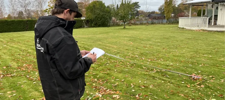 En person i mørk jakke og caps står på en gressplen og noterer på et ark mens et målebånd ligger strukket utover plenen. Det ligger høstløv spredt på gresset, og i bakgrunnen sees trær, hekk og et hvitt hus med veranda.